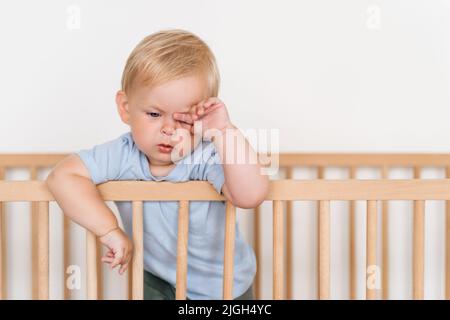 Ritratto di bello carino piccolo 11 mesi di età sonnolenta ragazzo stanco in abiti blu sfregamento occhi che si addormentano in piedi nel letto appoggiandosi sui paraurti isolato su sfondo bianco. È il momento di fare un pisolino Foto Stock