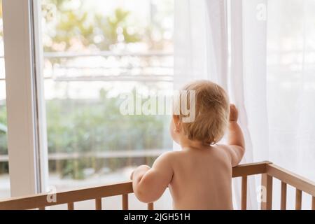 Vista posteriore del bambino solitario triste in piedi nel letto guardando attraverso la finestra in attesa di ritorno di sua mamma o papà, lasciato solo a casa. Trauma infantile. Cattive relazioni familiari. Assistenza all'infanzia Foto Stock