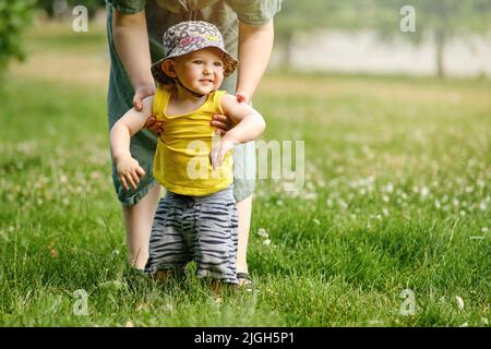 La madre insegna a camminare felice bambino bambino ragazzo in natura su erba verde. Bambino sorridente che prende i primi passi in estate parkwith mamma. Bambino di età un anno Foto Stock