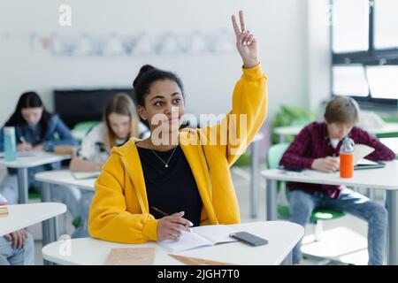 Gli studenti delle scuole superiori prestano attenzione in classe, seduti sulle loro scrivanie e alzando la mano. Foto Stock