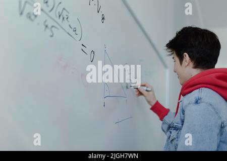 Vista posteriore dello studente delle scuole superiori che risolve il problema di matematica sulla lavagna bianca in aula. Foto Stock