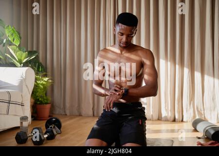 African American giovane maschio nero che si allenano a casa controllando l'orologio sportivo e in procinto di iniziare il suo esercizio sul tappeto yoga nel suo soggiorno Foto Stock