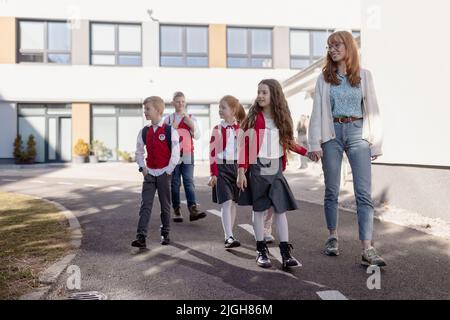 Ragazzi di scuola felici in uniformi con insegnante che cammina in un cortile. Foto Stock