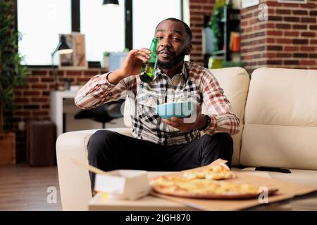 Ragazzo felice che beve birra alcolica dalla bottiglia e che tiene ciotola di patatine, guardando il film sul programma televisivo. Gustando una bevanda con consegna da asporto, cibo e spuntini, intrattenimento. Foto Stock