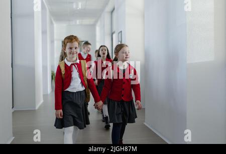 Felice schoolgrirl con sindrome di Down in uniforme tenuta mani con il suo compagno di classe a piedi in corridoio sciolto con compagni di classe dietro di loro. Foto Stock