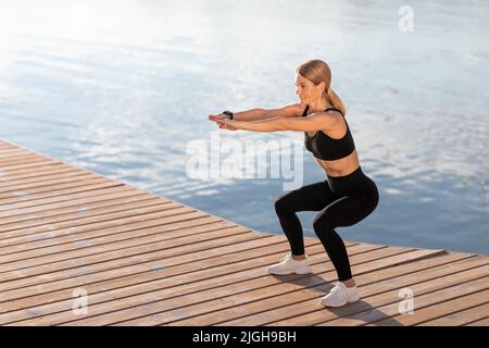 Formazione all'aperto. Sorridendo donna di mezza età facendo squats esercitarsi sul molo, Foto Stock