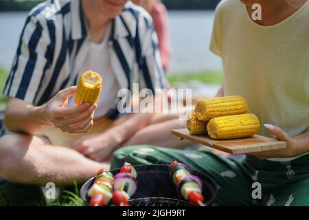 Primo piano di giovani amici che mettono il mais sulla griglia e che hanno barbecue quando si campeggio in campeggio. Foto Stock