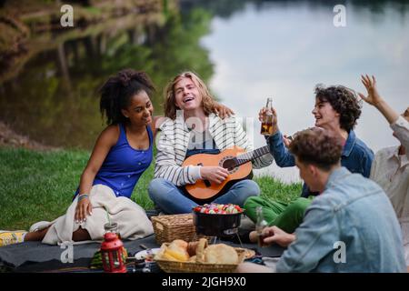 Gruppo di amici che si divertono al picnic vicino a un lago, seduti su coperta mangiare e suonare la chitarra. Foto Stock