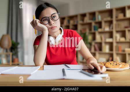 Noiosa ragazza asiatica teenage stanca in occhiali con cuffie studiare a distanza, fare una pausa, riposare Foto Stock