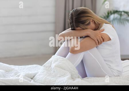 Donna bionda turbata seduta a letto a casa, piangendo Foto Stock