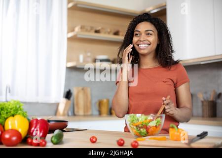 Allegra giovane afroamericana che prepara l'insalata con verdure biologiche parla per smartphone Foto Stock