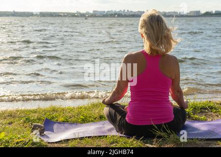 Una vista dal retro di una donna anziana seduta nella posa di Ardha Padmasana, una mezza posa di yoga di loto, su un tappeto nel parco e guardando il lago. SP Foto Stock
