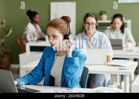 Operatore di assistenza clienti femminile in cuffie seduti al tavolo con un notebook e parlando online con i clienti al servizio di assistenza Foto Stock