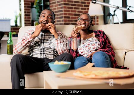 Giovane uomo e donna che mangiano fetta di pizza e bevono bottiglie di birra mentre guardano il film in tv insieme. Goditi la consegna rapida di cibo da asporto e bevande, guardando la televisione. Foto Stock
