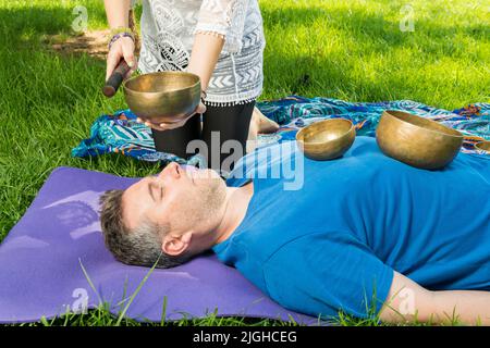 Un uomo sdraiato su un tappeto di yoga con ciotole cantanti sul suo corpo mentre una donna esegue una sessione di terapia musicale con ciotole cantanti su una zona erbosa. Foto Stock