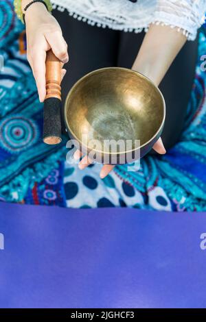 Vista dall'alto di una donna che tiene e suona una ciotola di canto tibetano durante una sessione di meditazione e di terapia musicale su un tappetino yoga. Foto Stock