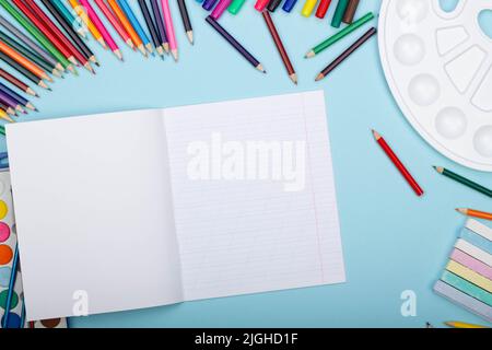 Foglio bianco di quaderno a linea obliqua, forniture per la pittura della scuola d'arte, su sfondo blu con spazio di copia per il testo. Matite colorate, pennarelli Foto Stock