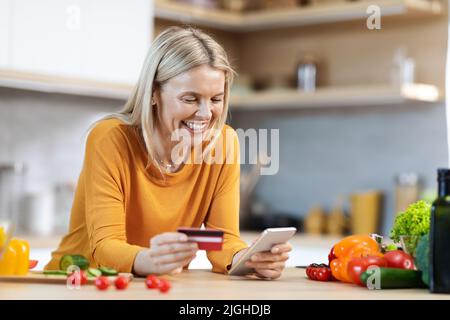 Donna bionda felice che acquista cibo online, utilizzando lo smartphone Foto Stock
