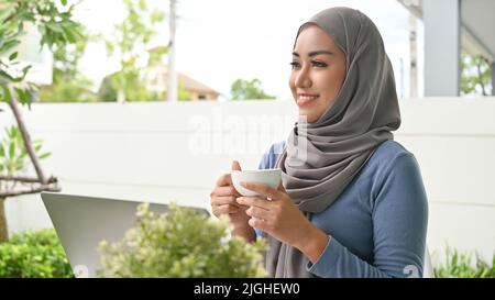 Bella donna musulmana asiatica millenaria con hijab rilassa sorseggiando un caffè del mattino nel suo cortile di casa. Foto Stock