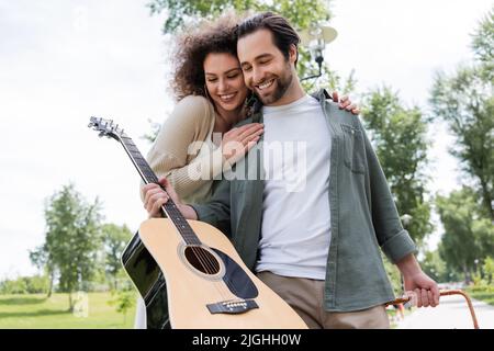 uomo sorridente in abiti estivi che tiene la chitarra vicino ragazza felice nel parco verde Foto Stock
