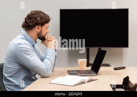 Vista laterale di un uomo d'affari maturo seduto alla scrivania, guardando un webinar o facendo videochiamate, utilizzando il computer, mockup Foto Stock