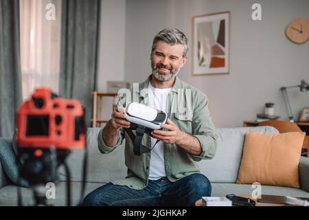 Happy europeo vecchio blogger maschio con barba mostra occhiali vr, fa video recensione in salotto interno Foto Stock