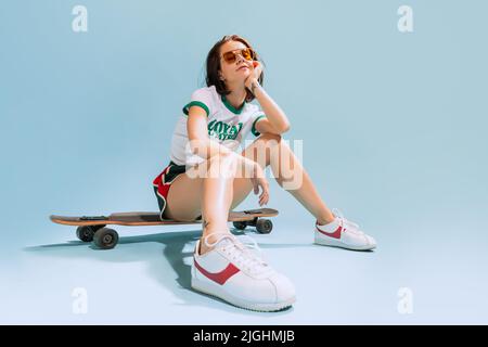 Ritratto di giovane donna elegante seduta sullo skateboard, in posa isolata su sfondo blu studio Foto Stock