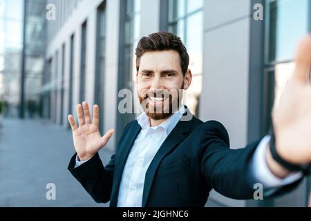 Felice fiducioso bel millenario europeo businessman bearded prende selfie, ondeggiando la mano sulla strada Foto Stock