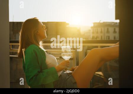 Donna con vino bianco gode il tramonto sul suo balcone. Foto Stock