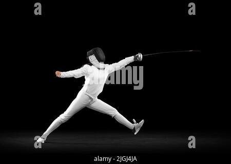 Fencer femminile energico in costume bianco e maschera in azione, movimento isolato su sfondo scuro. Sport, gioventù, attività, abilità, realizzazioni Foto Stock