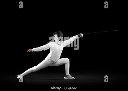 Fencer femminile energico in costume bianco e maschera in azione, movimento isolato su sfondo scuro. Sport, gioventù, attività, abilità, realizzazioni Foto Stock