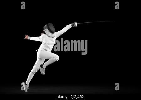 Fencer femminile energico in costume bianco e maschera in azione, movimento isolato su sfondo scuro. Sport, gioventù, attività, abilità, realizzazioni Foto Stock