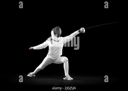 Fencer femminile energico in costume bianco e maschera in azione, movimento isolato su sfondo scuro. Sport, gioventù, attività, abilità, realizzazioni Foto Stock