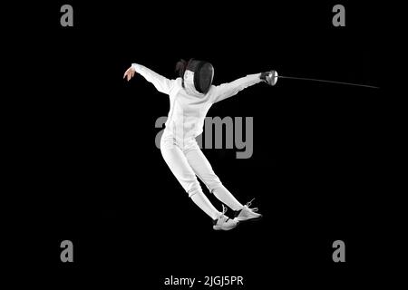 Fencer femminile energico in costume bianco e maschera in azione, movimento isolato su sfondo scuro. Sport, gioventù, attività, abilità, realizzazioni Foto Stock