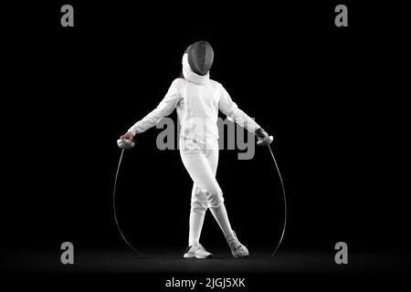 Giovane ragazza seria, fencer principiante in costume di scherma bianco in piedi con la maschera di fans di rapier isolato su sfondo scuro. Sport, giovani, successi Foto Stock