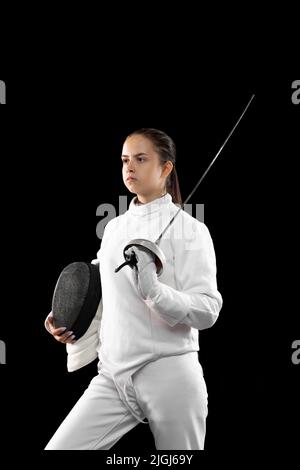 Giovane ragazza seria, fencer principiante in costume di scherma bianco in piedi con la maschera di fans di rapier isolato su sfondo scuro. Sport, giovani, successi Foto Stock