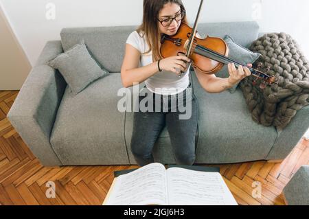 vista dall'alto, giovane donna venezuelana caucasica latina, con occhiali concentrati a suonare il violino seduto sul divano a casa. pratica classica Foto Stock