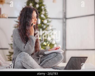 Giovane bella donna caucasica con capelli scuri in morbido maglione a maglia grigio usando il laptop sul letto in camera con albero di Natale. Decide le questioni relative alle prossime vacanze, ordini di consegna, effettua acquisti Foto Stock