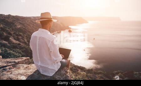 Nomade digitale, freelance. Lavoro remoto in vacanza. L'uomo nel cappello, un uomo d'affari con un computer portatile si siede sulle rocce vicino al mare durante il tramonto, fa un Foto Stock