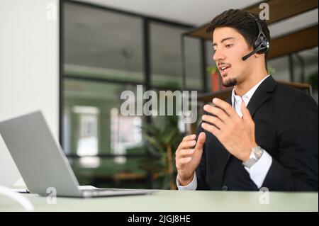 Un dipendente ispanico maschio, disponibile e cordiale, che indossa il dispositivo auricolare, si trova di fronte al laptop in ufficio, guarda la webcam e parla a distanza, uomo d'affari che utilizza le cuffie con microfono Foto Stock