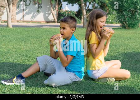 Bambini gli scolari mangiano gli hamburger nel parco seduti sull'erba. Pranzo scolastico. Foto Stock