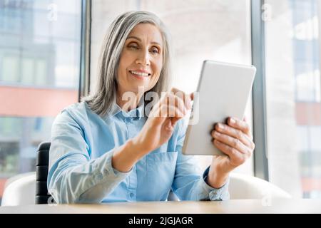 60s donne mature più anziane si siedono in ufficio e usano un tablet digitale Foto Stock