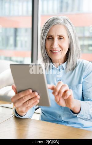 Ritratto verticale di una vecchia donna matura seduta in ufficio e utilizzare tablet digitale Foto Stock