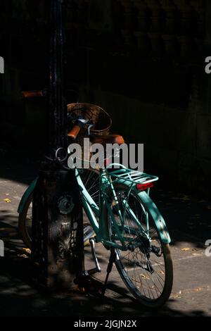 Bicicletta turchese con un cesto anteriore incatenato ad un lampione sotto l'ombra di un albero. Foto Stock