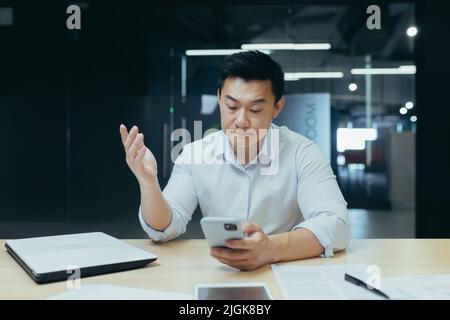 Turbato l'uomo d'affari asiatico che lavora in ufficio, leggendo cattive notizie al telefono, uomo depresso al lavoro Foto Stock