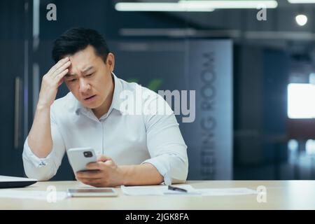 Turbato l'uomo d'affari asiatico che lavora in ufficio, leggendo cattive notizie al telefono, uomo depresso al lavoro Foto Stock