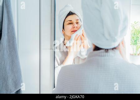 Donna in accappatoio con un asciugamano sulla testa guardando nello specchio e asciugando il viso con un asciugamano in bagno. Il concetto di procedure cosmetiche per la pelle Foto Stock