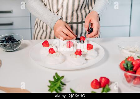 Cucina, facendo meringa fatta in casa. Donne mani che decorano torte Pavlova con frutti di bosco freschi sul tavolo da cucina con ingredienti. Ricette per delicio Foto Stock