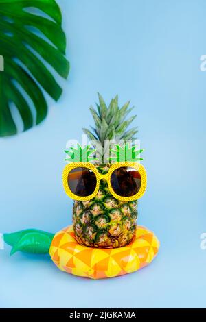 Ananas divertente in un bicchiere di ananas forma sul giallo gonfiabile anello su sfondo blu verticale. Estate in stile tropicale sulle vibrazioni della spiaggia. Foto Stock