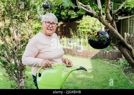 Ritratto di felice, sorridente anziano, donna matura innaffiare fiori con annaffiatura in giardino in una giornata di sole. Donna anziana in pensione e giardinaggio h Foto Stock